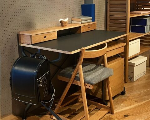 Wooden desk with a black top and small drawers, chair, and a backpack beneath on a pegboard wall workspace.
